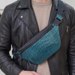 May include: Teal and black leather bum bag with a textured crocodile pattern. The bag has a black zip and strap, and a silver-coloured buckle. The bag is worn across the body.