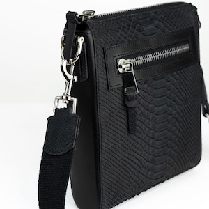 Pode incluir: Bolsa transversal preta com um padrão texturizado semelhante à pele de cobra. Possui zíper prateado, um bolso frontal e uma alça preta removível. A bolsa tem formato retangular.
