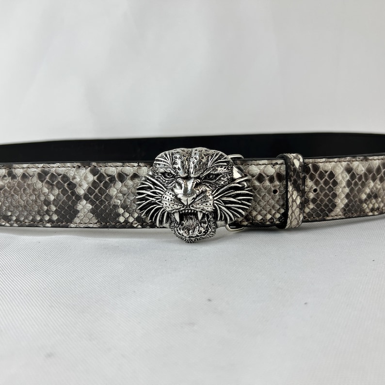 Pode incluir: Um cinto com padr&atilde;o de pele de cobra em tons de cinza e branco. O cinto apresenta uma fivela prateada em forma de cabe&ccedil;a de tigre rugindo. Um acess&oacute;rio elegante.
