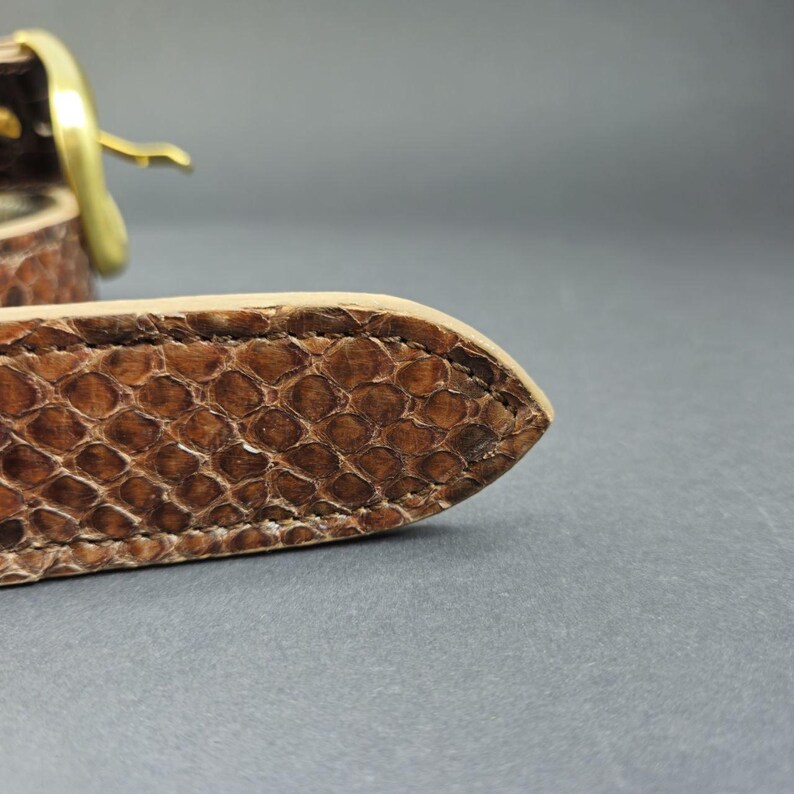 K&ouml;nnte beinhalten: Ein brauner G&uuml;rtel mit texturiertem Schlangenledermuster, spitz zulaufendem Ende und sichtbaren N&auml;hten an den Kanten. Der G&uuml;rtel wird mit einer polierten, goldfarbenen Metallschnalle geschlossen, die eine geschwungene Form aufweist.