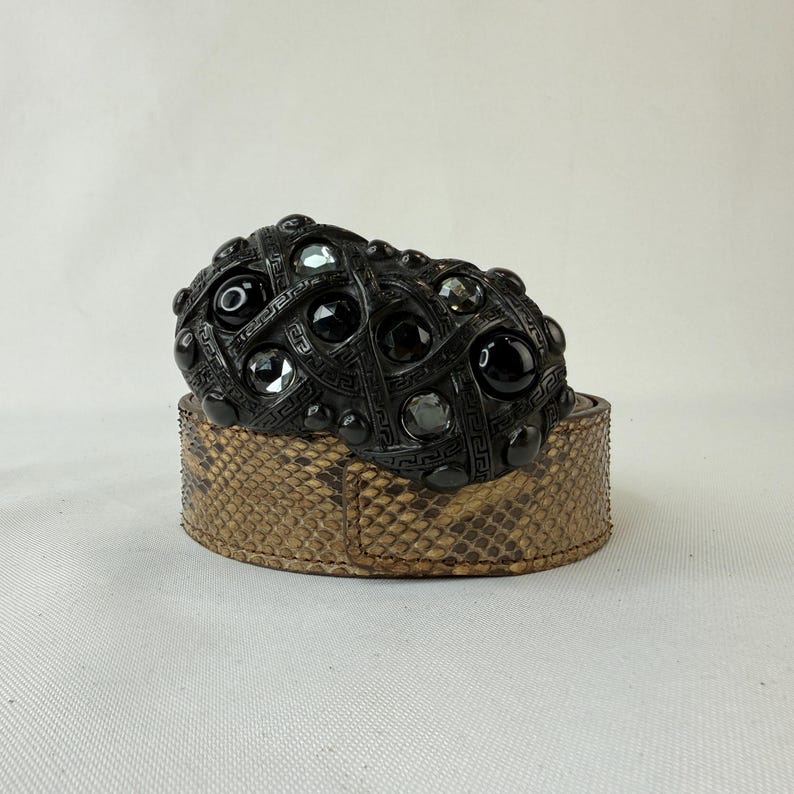 May include: A black belt buckle with a geometric design and embedded black and clear stones sits atop a snakeskin-patterned belt. The buckle has a woven pattern and is a dark, matte black color.