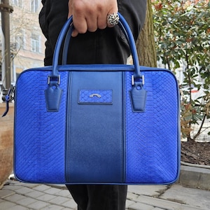 May include: A blue leather briefcase with a snakeskin texture on the sides and a smooth central panel. It features a top handle, silver hardware, and a silver emblem. The bag is a bright shade of blue, with a classic design.