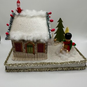 Puede incluir: Una escena de un pueblo navideño en miniatura con una casa de piedra, un tejado cubierto de nieve y detalles rojos. Una pequeña figura roja está en el tejado, otra esquiando cerca. La base está adornada con oropel verde y dorado.