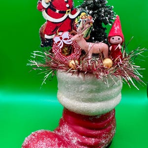 Könnte beinhalten: Festliche Weihnachtsdekoration mit einem roten Stiefel mit weißem Bund, verziert mit Miniaturfiguren. Weihnachtsmann, Rentier und ein kleiner Weihnachtsbaum gehören zu den Dekorationen. Der Stiefel ist mit Glitzer verziert, was einen funkelnden Effekt erzeugt.
