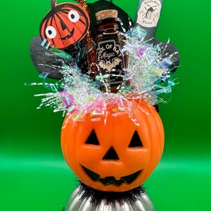 Puede incluir: Decoración temática de Halloween con una linterna de calabaza naranja sobre una base plateada con oropel negro. Incluye un murciélago, un recorte de calabaza y botellas etiquetadas como "Pool of Bat Magic" y "Beetle Juice Elixir".