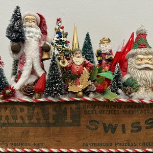 Könnte beinhalten: Ein Vintage-Weihnachtsdiorama mit Weihnachtsmannfiguren, Miniatur-Tannenbäumen und dekorativen Elementen, das in einer Holzkiste von Kraft Swiss präsentiert wird. Die Kiste ist mit einem rot-weißen Seil verziert.