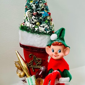 Può includere: Una decorazione natalizia vintage con un piccolo albero decorato, una calza rossa con la scritta "Merry Christmas" e una statuina di elfo. L'albero è adornato con ornamenti e neve, e l'elfo indossa un cappello verde e un abito rosso.