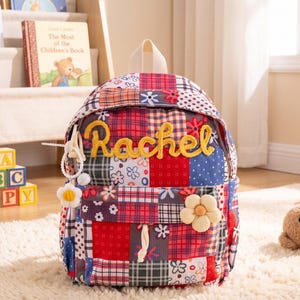 May include: A patchwork backpack featuring a colorful mix of red, blue, and white plaid and floral patterns. The front displays the name Rachel in yellow cursive lettering, a white flower charm, and a large, plush, cream-colored flower applique.