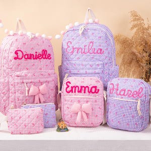 May include: Two backpacks, two crossbody bags, and one small pouch in pink and purple gingham fabric with quilted textures. The bags feature embroidered names Danielle, Emilia, Emma, and Hazel, along with decorative fabric bows on the front.