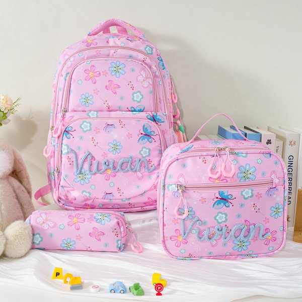 Könnte beinhalten: Ein Set aus einem Rucksack, einer Lunchtasche und einem Federmäppchen in Rosa mit einem Muster aus blauen und rosa Blumen und Schmetterlingen. Jeder Artikel ist mit dem Namen Vivian in blauer Schreibschrift auf der Vorderseite bestickt.