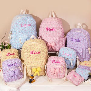 May include: A collection of quilted backpacks, crossbody bags, and pouches in various pastel colors, including blue, pink, yellow, and purple. Each bag features a gingham pattern and is embroidered with a name. Some bags have bow accents.