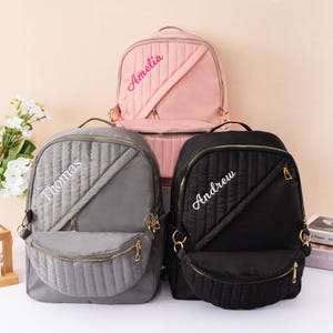 May include: Three personalized backpacks in pink, gray, and black. Each backpack features a quilted design, a front zippered pocket, and a gold zipper. The names "Amelia", "Thomas", and "Andrew" are embroidered on the front.