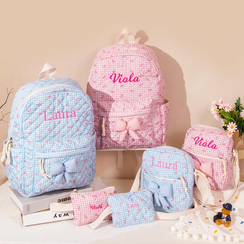 May include: A collection of personalized backpacks and matching accessories in pink and blue gingham patterns. The backpacks feature embroidered names and bow accents. Includes a backpack, small purse, and a small pouch.