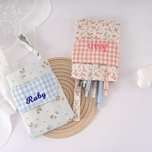 May include: Two fabric pencil cases with floral and gingham patterns. One is light blue with "Ruby" embroidered in blue, the other is floral with "Jean" embroidered in pink. Pens and pencils are visible in the floral case.