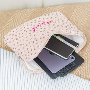 May include: A pink floral quilted laptop sleeve with a zipper, containing a tablet with a keyboard case, a notebook, and a pen. The word "marcel" is embroidered in pink on the sleeve.