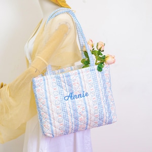 May include: A quilted tote bag with blue and white floral and gingham patterns. The bag has a light blue handle and the name "Annie" embroidered in a matching blue thread. The bag is filled with pink roses.