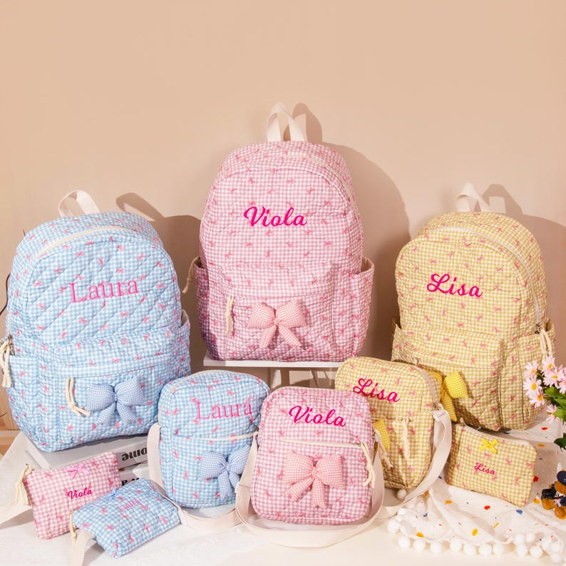 May include: A collection of personalized backpacks, purses, and pouches in various sizes and colors. The bags feature a gingham pattern in blue, pink, and yellow, with embroidered names and bow accents. The backpacks have a height of approximately 12 inches.