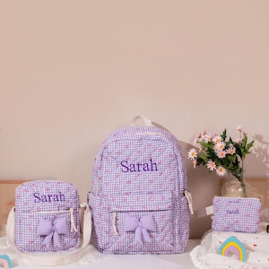May include: A matching set of three items: a backpack, a crossbody bag, and a small pouch. All items are made of a lavender and white gingham fabric with embroidered text that reads "Sarah". Each item has a lavender bow.
