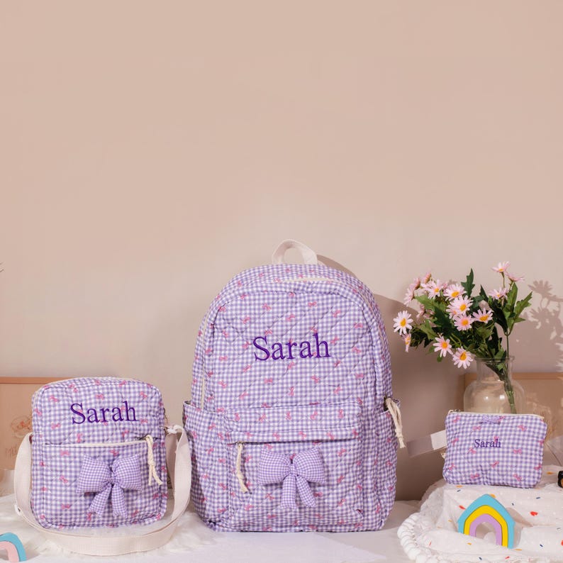May include: A matching set of three items: a backpack, a crossbody bag, and a small pouch. All items are made of a lavender and white gingham fabric with embroidered text that reads "Sarah". Each item has a lavender bow.
