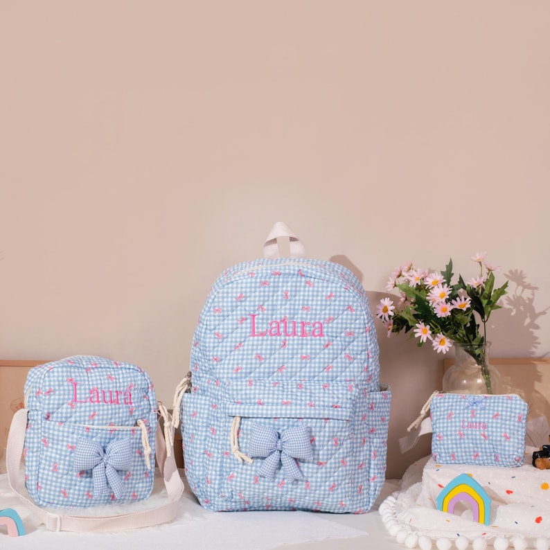May include: A set of three matching blue gingham bags: a backpack, a crossbody bag, and a small pouch. Each bag features a pink embroidered name and a bow. The backpack has a front pocket and adjustable straps.