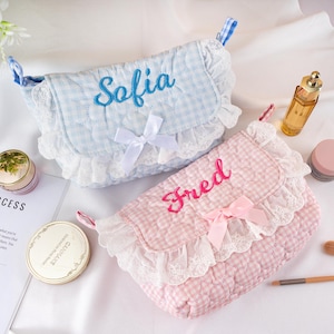 May include: Two quilted cosmetic bags, one light blue and one pink, with gingham patterns. Each bag features a name embroidered on the flap, Sofia and Fred, and is trimmed with white lace and a bow. The bags are designed for storing makeup and beauty products.