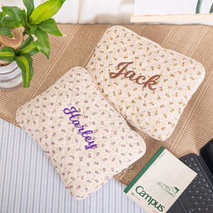 May include: Two quilted pillows with floral patterns and embroidered names. One pillow has the name "Jack" in brown thread, and the other has "Harley" in purple thread. The pillows are cream-colored with small yellow and purple flowers.
