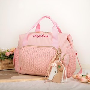 May include: Pink diaper bag with the name "Sophia" embroidered on the front. The bag has a braided texture on the front pocket and includes a matching keychain with a horse design. Features a top handle and shoulder strap.