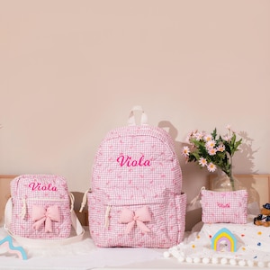 May include: A set of pink gingham backpacks and accessories. The set includes a backpack, a crossbody bag, and a small pouch, all with the name "Viola" embroidered in pink. Each item features a pink bow.