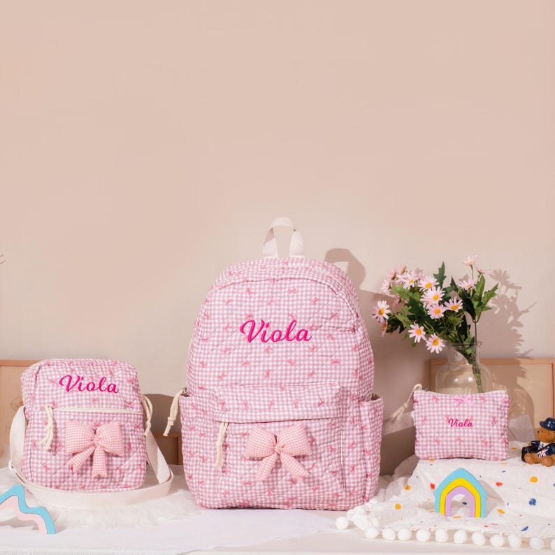 May include: A set of pink gingham backpacks and accessories. The set includes a backpack, a crossbody bag, and a small pouch, all with the name "Viola" embroidered in pink. Each item features a pink bow.