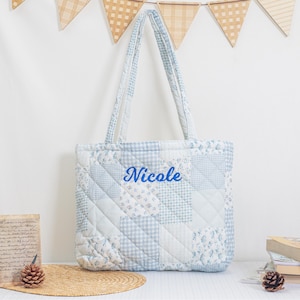 May include: A light blue quilted tote bag with a patchwork design. The bag features a variety of light blue patterns, including floral and gingham. The name "Nicole" is embroidered in blue on the front. The bag has two handles.