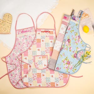 May include: A collection of colorful floral aprons with matching oven mitts. The aprons feature patchwork designs, floral patterns, and the names "Bonnie" and "Aurora". The aprons are in shades of pink, blue, and yellow.