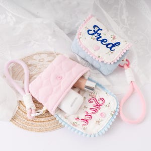 May include: Two small, quilted fabric pouches. One is light pink and open, revealing a tube of makeup and a white object. The other is light blue with the name "Fred" embroidered in blue. Both have floral patterns and pink accents.