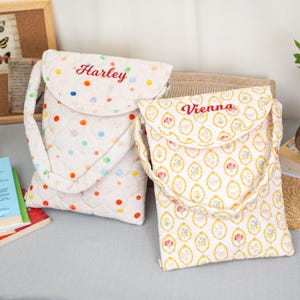 May include: Two quilted fabric crossbody bags. One bag is white with colorful polka dots and the name "Harley" embroidered in red. The other bag is white with a floral pattern and the name "Vienna" embroidered in red.