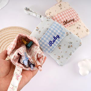 May include: A collection of small, floral-patterned fabric pouches in shades of pink, blue, and cream. One pouch is open, revealing pens and clips. Two other pouches have embroidered names, "Mary" and "Ruby", on gingham patches. Each pouch has a loop and button closure.