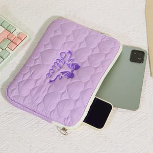 May include: A lavender quilted tablet sleeve with a heart pattern. The sleeve has a zipper closure and features the name "Alice" embroidered in purple script with a bow design. A tablet and a phone are partially visible.
