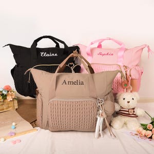 May include: A collection of personalized baby bags in black, pink, and tan. Each bag features a name embroidered in a contrasting color. The tan bag has a textured front pocket. A teddy bear and flowers are also in the image.