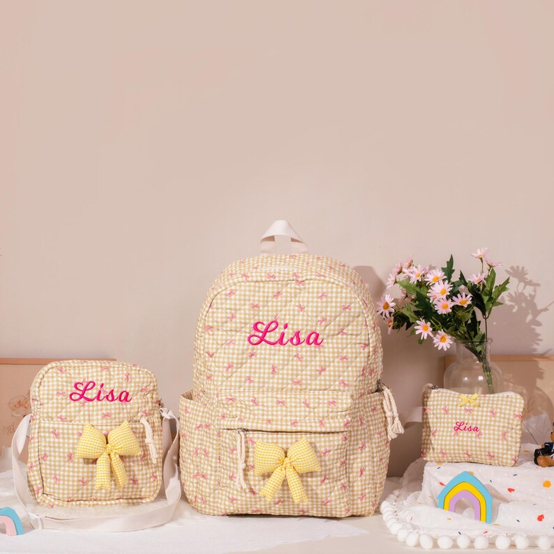 May include: A set of three matching bags: a backpack, a crossbody bag, and a small pouch. All bags are beige with a gingham pattern and pink embroidered text that reads "Lisa". Each bag has a yellow bow.