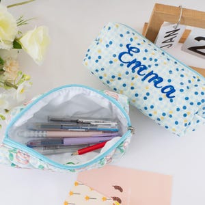 May include: Two fabric pencil cases. One is open, revealing pens and pencils. The other is closed, with the name "Emma" embroidered in blue. Both cases have a light blue and white polka dot pattern. The open case has a floral pattern.