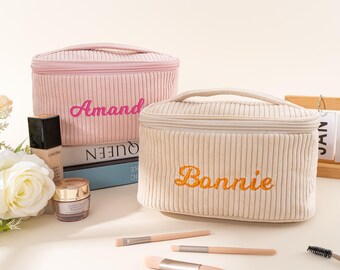 May include: Two personalized makeup bags, one pink with the name "Amanda" embroidered in pink and one beige with the name "Bonnie" embroidered in orange. The bags are made of a soft, ribbed fabric.