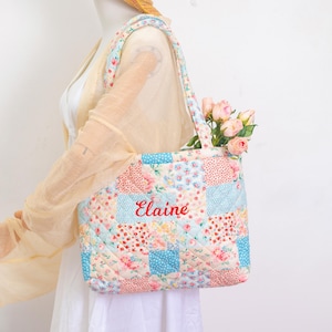 May include: A quilted patchwork tote bag with floral and polka dot patterns in pastel colors. The bag features the name "Elaine" embroidered in red. The bag is filled with pink roses and has matching quilted handles.