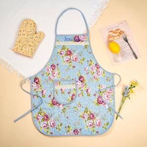May include: A light blue quilted apron with a floral pattern of pink and red roses. The apron has a pocket and is trimmed with lace and a blue polka dot border. The name "Jessica" is embroidered on the bib. A matching oven mitt and a vintage recipe are also visible.