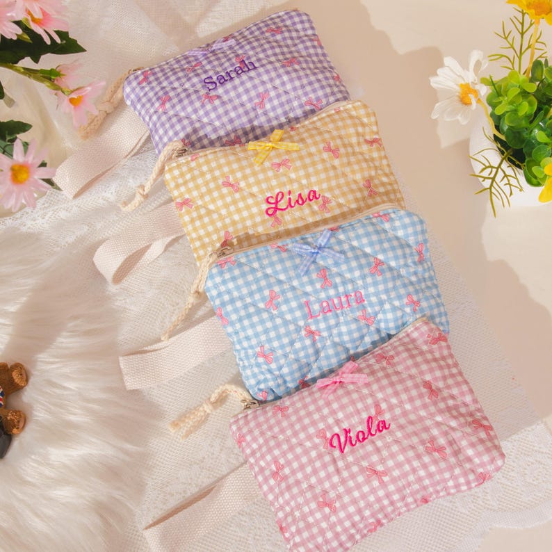 May include: Four quilted cosmetic bags in pastel colors: purple, yellow, blue, and pink. Each bag features a gingham pattern, embroidered name, and bow accent. The bags have a zipper closure and a cream-colored wrist strap.