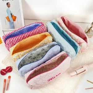 May include: A collection of open, rectangular pencil cases in various patterns and colors. The cases feature gingham, floral, and quilted designs. One case has the name "Flora" embroidered on it. The interiors of the cases are visible, showcasing different fabric patterns.