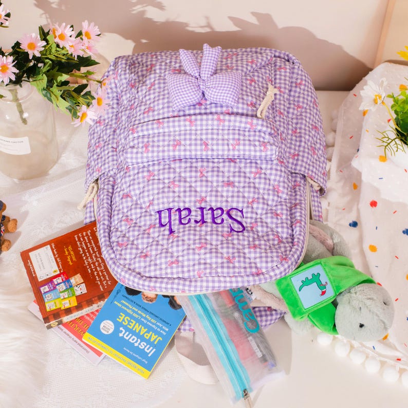 May include: A quilted purple backpack with a gingham pattern and the name "Sarah" embroidered in purple. The backpack has a bow detail and is open, revealing books, pens, and a plush turtle.