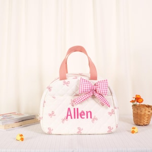 May include: A white quilted lunch bag featuring a repeating pattern of small pink bows. The bag has two pink fabric handles, a large pink gingham bow attached to the front, and the name Allen embroidered in pink script lettering on the center.