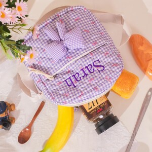 May include: A small, quilted backpack in a purple and white gingham pattern, featuring a bunny ear design and the name "Sarah" embroidered in purple. The bag is open, revealing a jar of coffee, a banana, and other items.