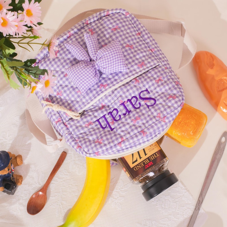 May include: A small, quilted backpack in a purple and white gingham pattern, featuring a bunny ear design and the name "Sarah" embroidered in purple. The bag is open, revealing a jar of coffee, a banana, and other items.