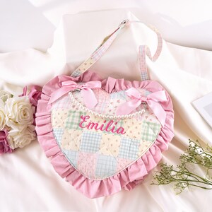 May include: A heart-shaped pink quilted bag with a ruffled edge and a floral and checkered pattern. The bag has a shoulder strap, pink bows, and the name "Emilia" embroidered in pink. A pearl necklace is attached to the bows.
