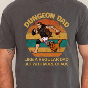 Puede incluir: Una camiseta gris carbón de manga corta con un gráfico de una persona corriendo y un gato dentro de un círculo de atardecer. El texto DUNGEON DAD LIKE A REGULAR DAD BUT WITH MORE CHAOS aparece en letras amarillas y blancas en la parte frontal.