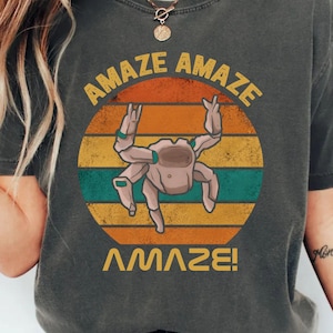May include: A charcoal gray short-sleeve t-shirt featuring a circular graphic with horizontal stripes in orange, yellow, and teal. The graphic depicts a light brown crab with the text AMAZE AMAZE arched above and the word AMAZE! printed below.